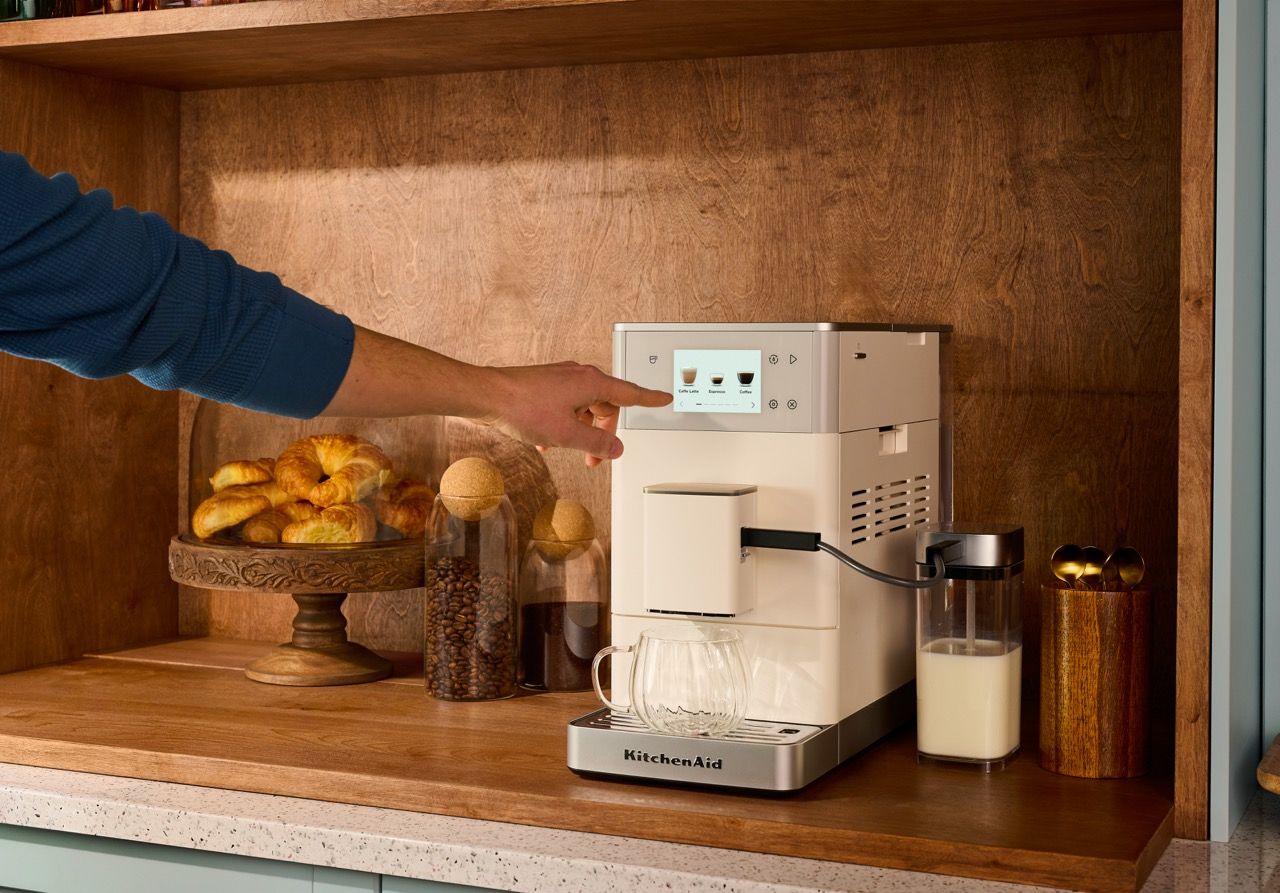 Hand tippt auf das Display des KitchenAid KF4 5KES8454EPL in Porcelain White; daneben Croissants, Bohnen im Glas und der integrierte Milchbehälter.