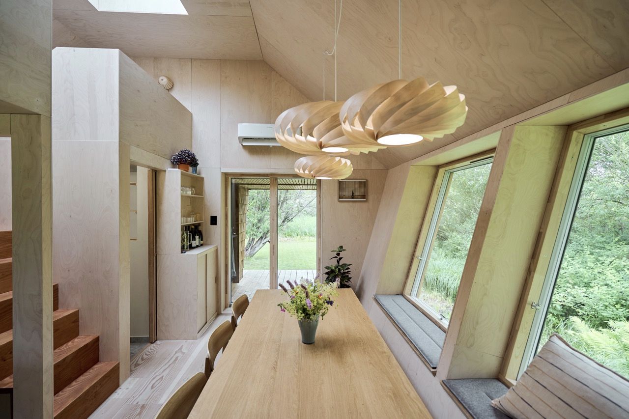 Essbereich mit langem Holztisch und Pendelleuchten aus Holzfurnier, schräg gestellte Fenster mit Sitznischen und Blick ins Grüne, oben ein Dachfenster.