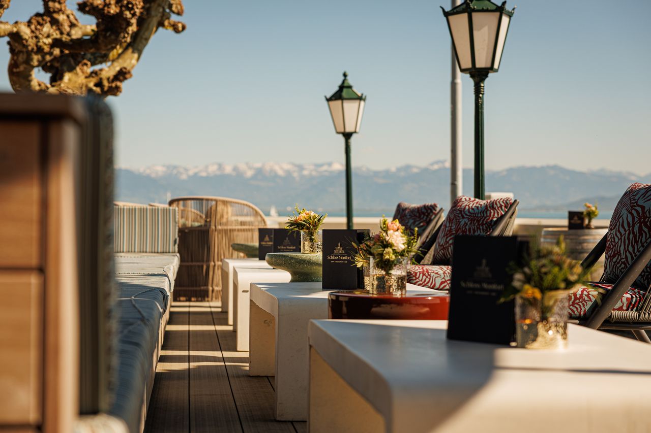 Sonnenterrasse des Schloss Montfort mit Sitzgelegenheiten, Blumenarrangements und Blick auf den Bodensee sowie die Alpen im Hintergrund.