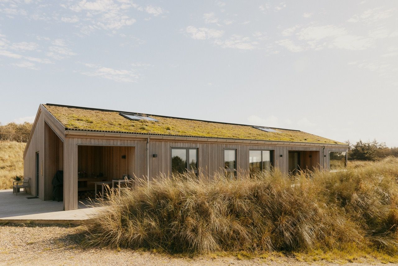 Langgestrecktes Ferienhaus mit Holzfassade und begr&uuml;ntem Dach mit Dachfenstern, umgeben von D&uuml;nengras unter hellem Himmel.