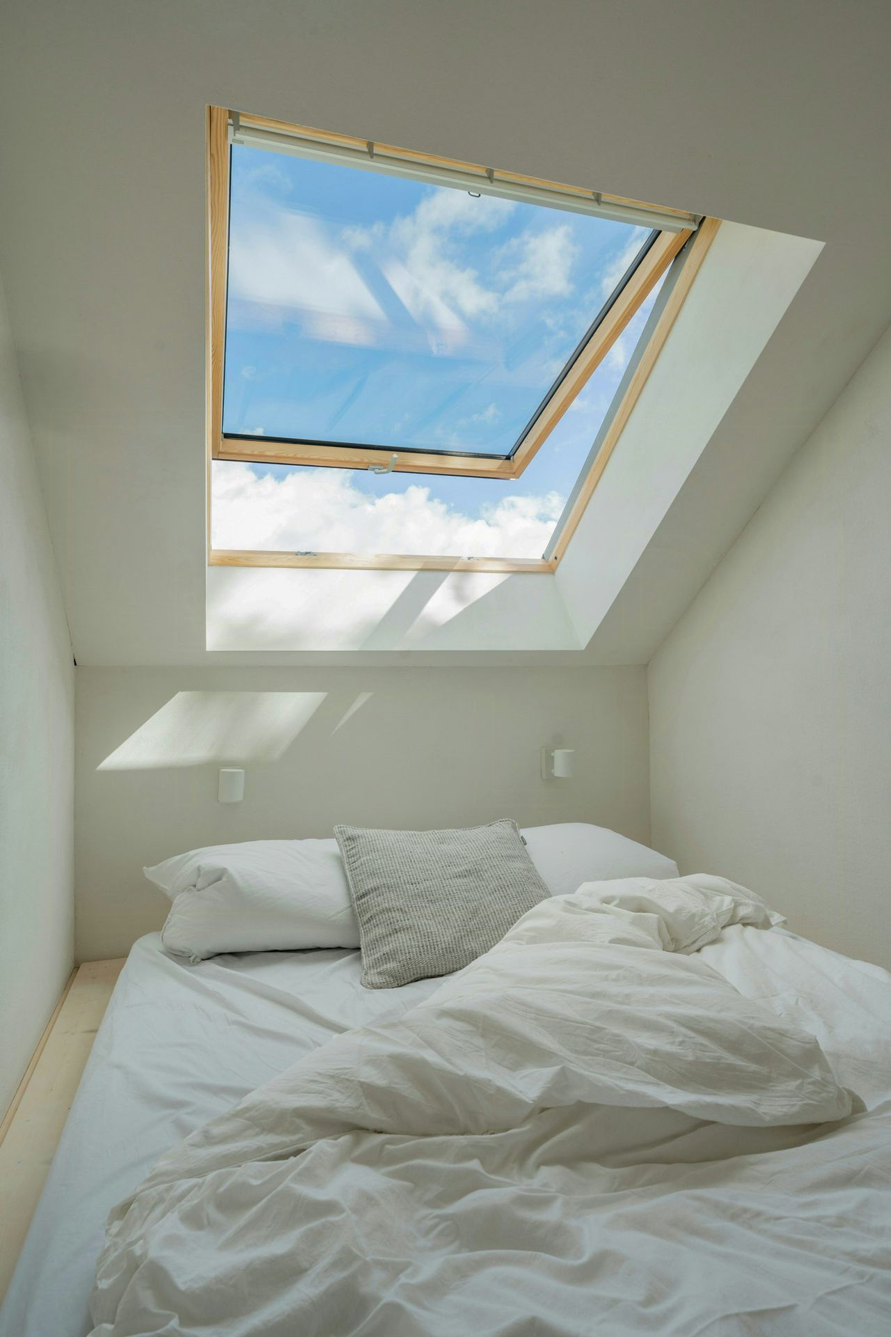 Weißes Schlafzimmer mit zerwühltem Bett, darüber ein geöffnetes VELUX Dachfenster mit Holzrahmen, durch das man blauen Himmel und Wolken sieht.
