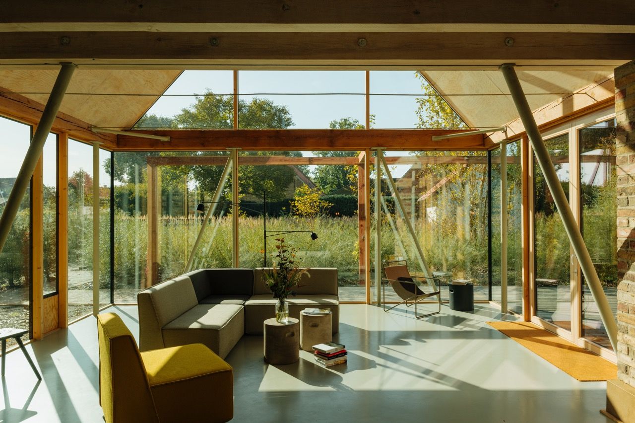 Wohnraum mit raumhohen Glaswänden, Holzbalken und schrägen Stützen, darin ein graues Ecksofa, ein gelber Sessel und Beistelltische auf hellem Betonboden. Rechts steht eine Backsteinwand, draußen wachsen hohe Gräser und Bäume im Sonnenlicht.