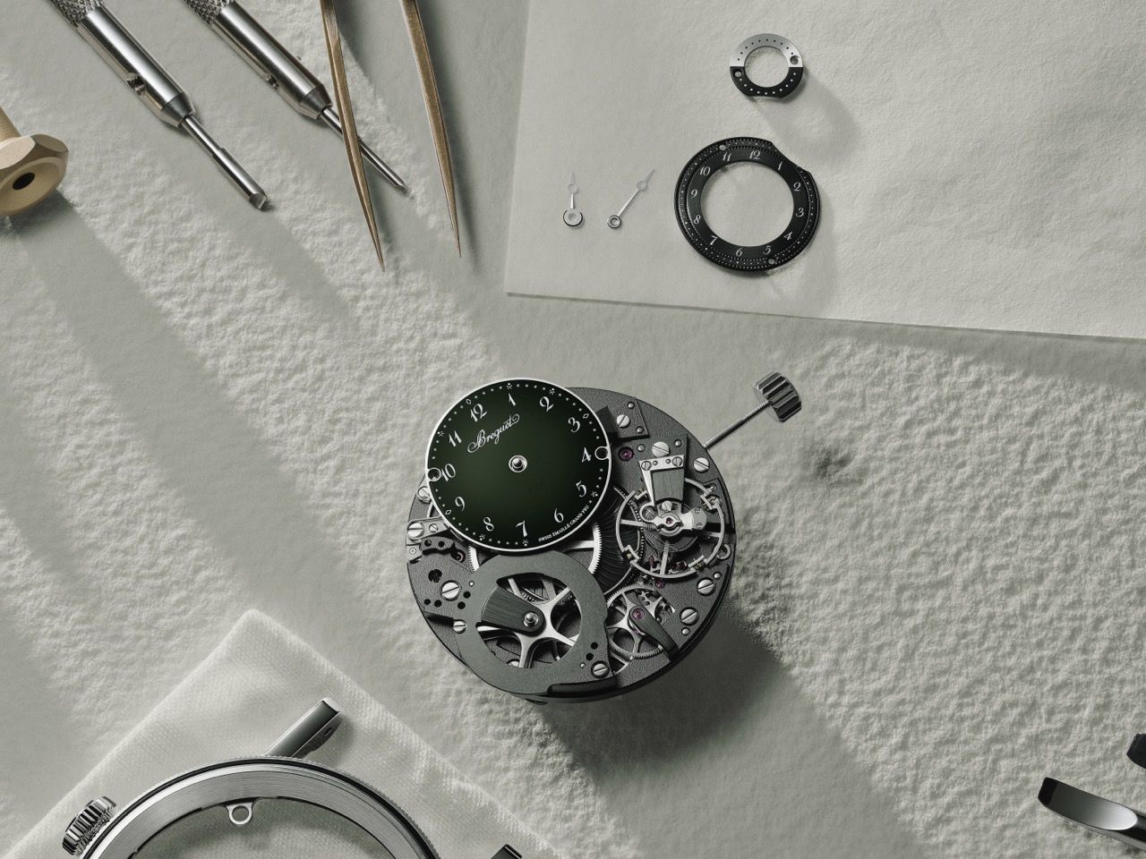Blick auf den Werkstatt-Arbeitstisch eines Uhrmachers bei der Montage einer Breguet Tradition Uhr mit Werkzeugen, Pinzetten und einzelnen Zifferblattkomponenten.