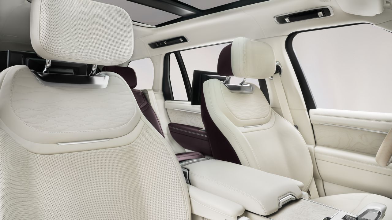 ine Innenansicht des luxuriösen Fond-Bereichs eines Range Rovers, der einen exklusiven „Bespoke“-Ansatz mit personalisierten Details zeigt. Das Bild ist aus der Perspektive des linken Rücksitzes aufgenommen und zeigt die beiden getrennten, verstellbaren Executive-Sitze. Die Sitze und die Mittelkonsole sind überwiegend in einem cremeweißen, perforierten und gesteppten Leder gehalten. Die Seitenverkleidungen der Sitze und die Türarmlehne sind in einem tiefen, reichen Violettton abgesetzt, was einen eleganten Zweifarben-Effekt erzeugt.
